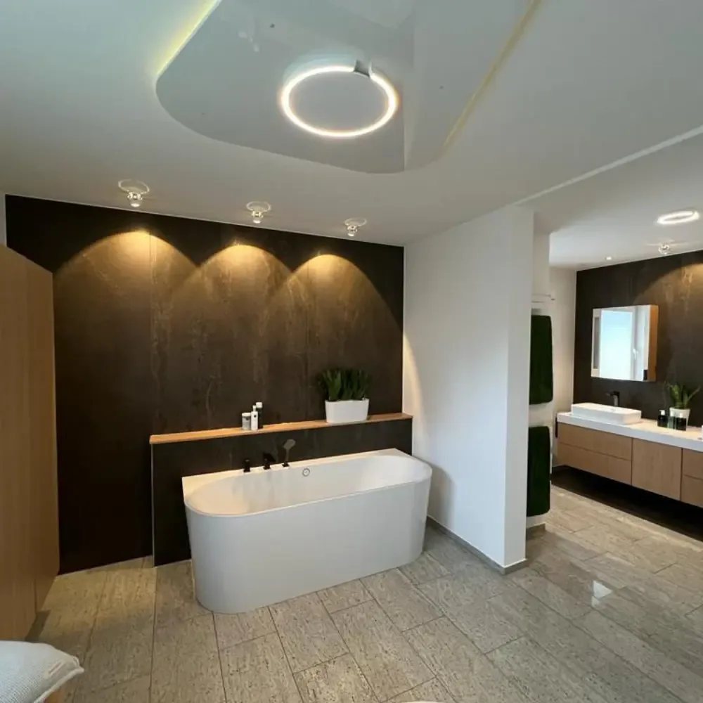 Elegantes Badezimmer mit freistehender Badewanne vor einer dunklen XXL-Keramik-Wandverkleidung in Natursteinoptik, warmem Akzentlicht, Eichenholz-Waschtisch und hellem Steinboden